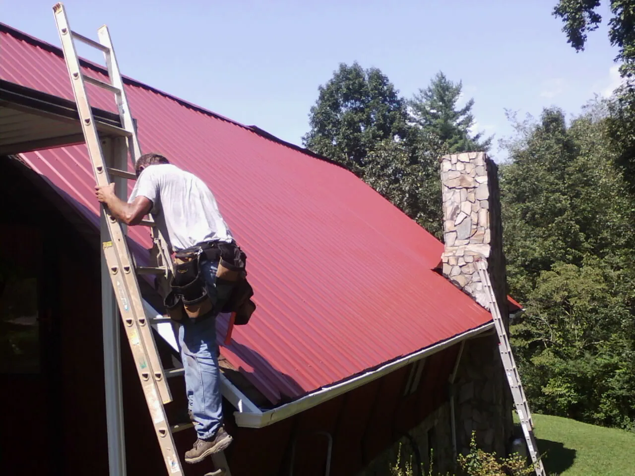 Roofer on a ladder beside a red metal roof and stone chimney — Soffit & Fascia Repair by Roofing Brothers in New Stanton